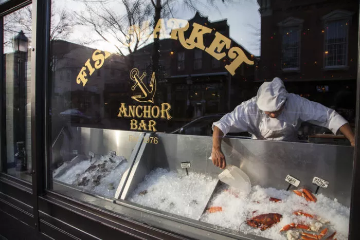 A fishmonger wearing a white uniform arranges ice and seafood in a display case inside a fish market, seen through a window with “Fish Market” and “Anchor Bar” written on it.