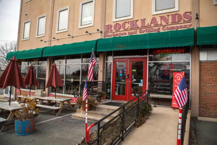 Exterior of Rocklands Barbeque and Grilling Company, featuring outdoor seating, American flags, a green awning, and a red door, with signage above the entrance.