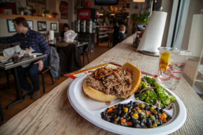 A plate with a pulled pork sandwich, black beans, and broccoli salad sits on a wooden counter in a casual restaurant. A cup with a straw is next to the plate, and diners are seated in the background.