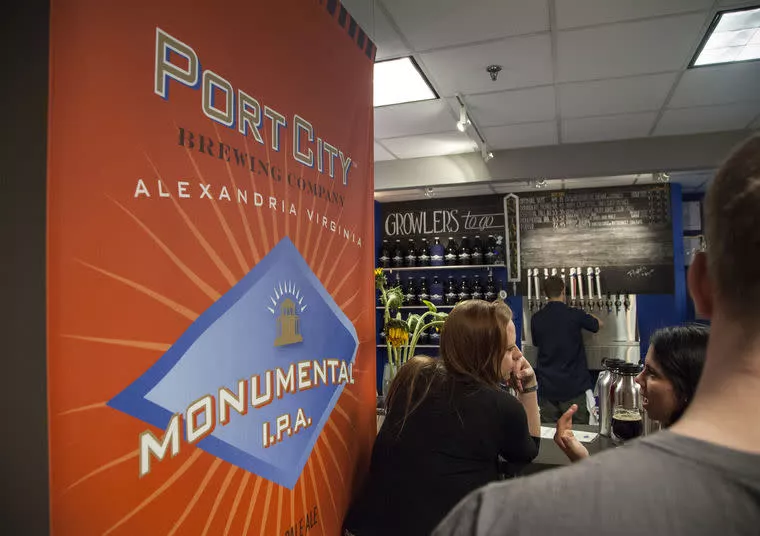 People stand at a bar with a large red-orange sign for Port City Brewing Company’s Monumental I.P.A. in the foreground. Behind the bar, a chalkboard beer list and growlers are visible on shelves.