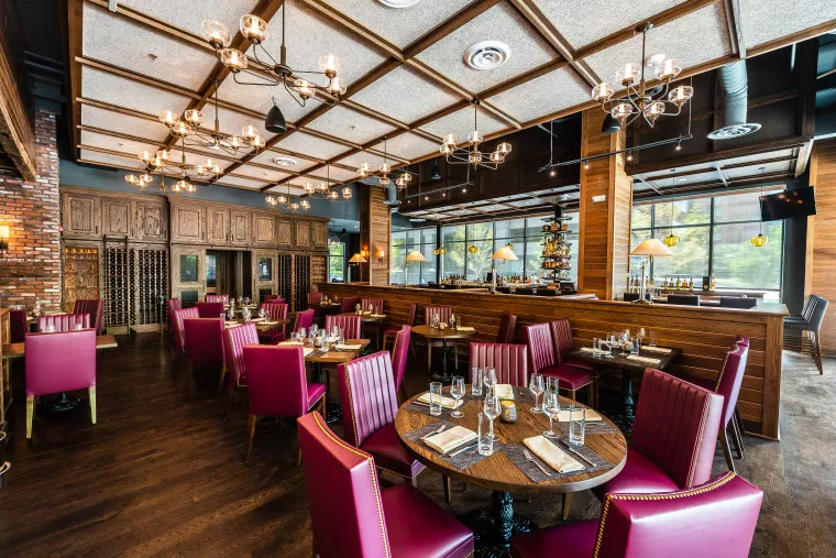 A stylish restaurant interior with burgundy chairs, wooden tables set for dining, a wooden paneled ceiling, modern chandeliers, large windows, and a bar area in the background.
