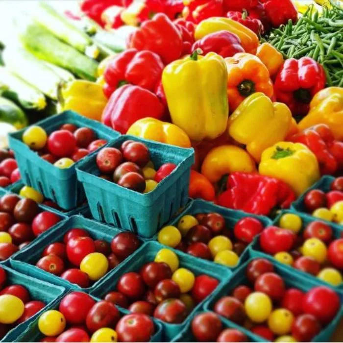 A colorful assortment of fresh vegetables, including cherry tomatoes in blue baskets, yellow and red bell peppers, green beans, and zucchini, displayed at a farmers market.