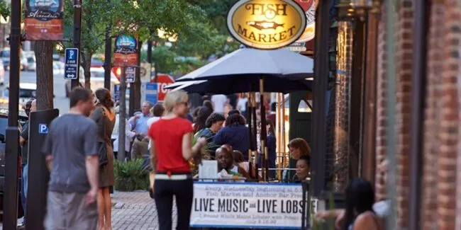 People walk and dine outside a bustling restaurant called Fish Market. Tables with umbrellas line the sidewalk, and a sign advertises live music and live lobster. Trees and street signs are visible along the brick-paved street.