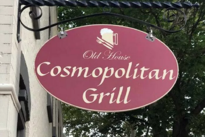 A red oval sign with cream-colored text reads Old House Cosmopolitan Grill, with a graphic of a beer mug and knife and fork above the text. The sign hangs outside, with trees in the background.