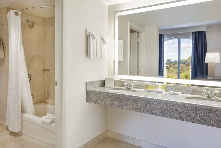 Modern hotel bathroom with a granite double sink, large illuminated mirror, toiletries, and a bathtub with a shower curtain. A window with blue curtains and a scenic outdoor view is visible in the adjacent room.