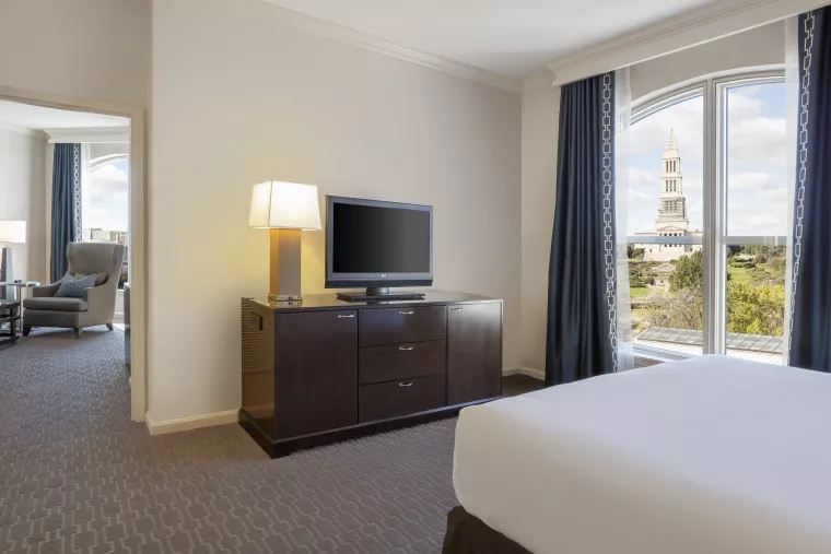 A modern hotel room with a bed, dresser with a TV, a lamp, and large windows showing a cityscape with a tall, tower-like building outside. An armchair is visible in an adjoining room.