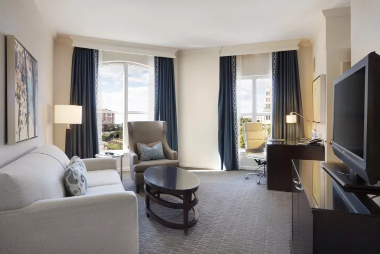 A bright hotel room with a sofa, armchair, coffee table, and flat-screen TV. Large windows with dark curtains reveal a city view. A desk with a chair and lamp is positioned near the windows. The decor is modern and neutral-toned.