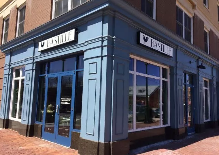 A corner view of a blue-painted building with large windows and double doors, displaying two signs that read “Bastille” with a rooster icon above the entrances.