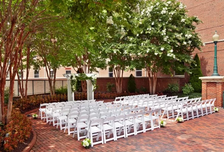 An outdoor wedding setup with rows of white chairs facing a white floral archway on a brick patio, surrounded by greenery and trees, next to a brick building and a lamppost.
