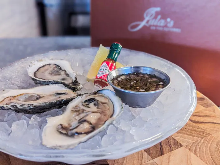 A plate of three raw oysters on ice, served with a lemon wedge, a small bottle of Tabasco sauce, and a metal cup of mignonette sauce, all on a wooden table. A restaurant menu is in the background.