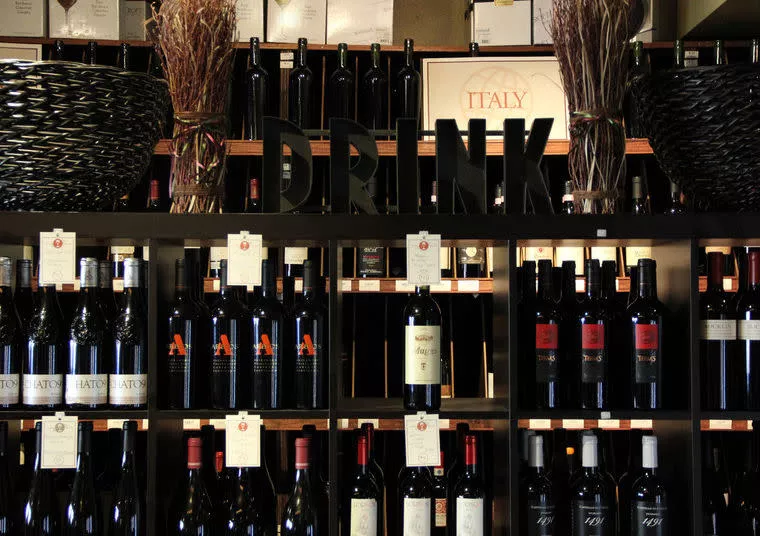 Shelves filled with various bottles of wine, organized in rows. A decorative sign spelling DRINK sits among the bottles, and baskets with twigs are placed at the top corners of the display.