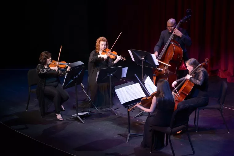 Five musicians dressed in black perform on stage, playing string instruments including violins, a cello, and a double bass. Music stands with sheet music are in front of them, and a red curtain is in the background.