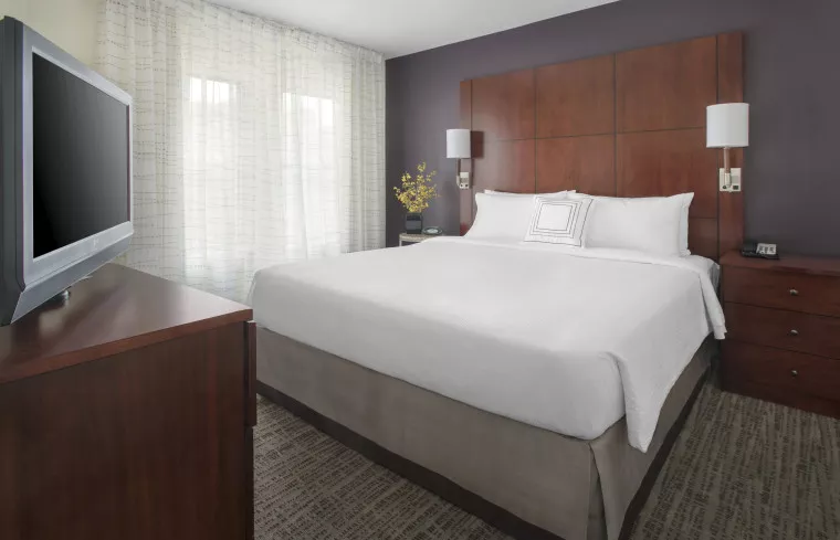 A modern hotel room with a large bed, white linens, wooden headboard, two bedside tables with lamps, a flat-screen TV on a wooden dresser, and sheer curtains over two windows.