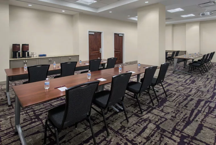 A conference room with rows of tables and chairs, each table set with bottled water and notepads. There is a refreshment station with coffee urns and cups at the back, and two closed double doors on the rear wall.