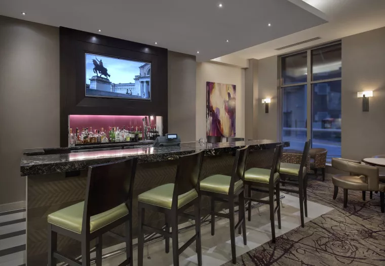 A modern hotel bar with five green padded chairs at a dark marble counter, shelves of liquor bottles behind, contemporary artwork on the wall, and a TV displaying a statue above the bar area.