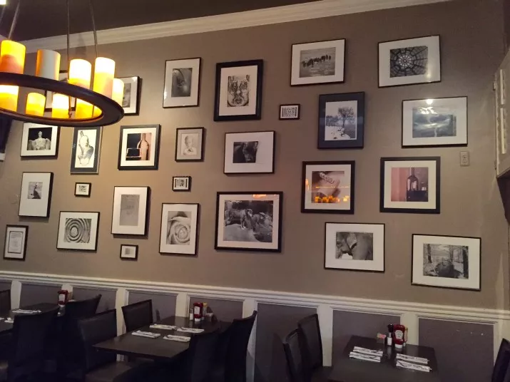 A restaurant wall displays a variety of framed black-and-white photos and artwork in different sizes above dining tables set with condiments and napkins. A modern chandelier with yellow lights hangs above.