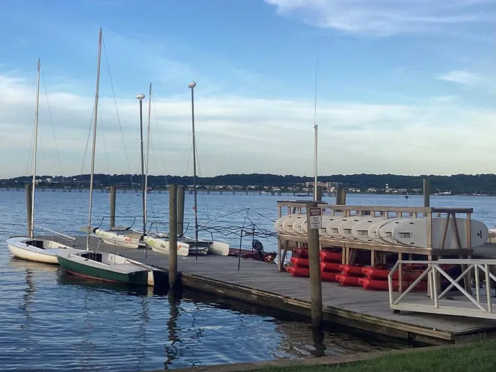 A wooden dock on a calm lake holds several small sailboats and stacked white canoes. Red life vests are stored under the canoes. Trees and a bridge are visible across the water under a blue sky with light clouds.