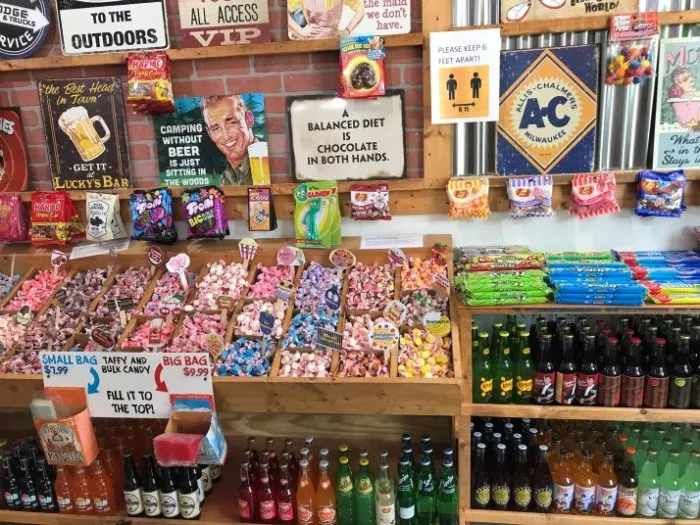 A colorful candy and soda shop display with bins of assorted candies, vintage-style signs, novelty chocolates, and glass bottles of soda. Signs encourage social distancing and promote various humorous messages.