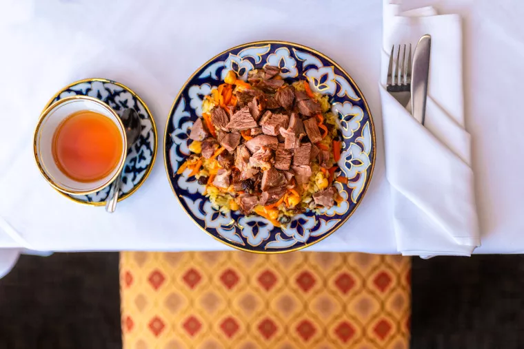 A plate of rice pilaf topped with chunks of meat sits next to a cup of tea with a spoon on a white tablecloth. A napkin with a fork and knife is placed beside the plate. A patterned chair is visible below the table.