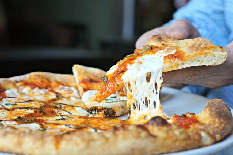 A hand lifts a cheesy slice of pizza from a whole pizza on a plate, showing melted cheese stretching from the slice.