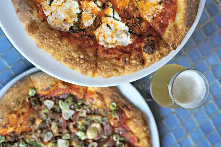Two plates of pizza, one with ricotta cheese and basil, the other topped with sliced sausages and scallions, are set on a blue tiled table next to a glass of beer.