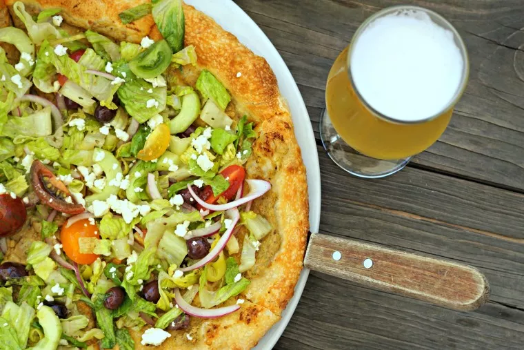 Closeup of a fresh veggie pizza topped with lettuce, tomatoes, red onions, olives, pepperoncini, and feta cheese on a wooden table next to a glass of light beer.