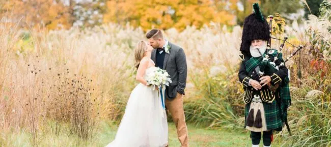 A bride and groom share a kiss outdoors in a field with autumn trees behind them, while a bagpiper in traditional Scottish attire plays nearby.