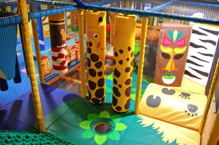 Indoor soft play area with jungle-themed padded equipment, including hanging pillars designed like giraffes, a zebra-patterned mat, and a colorful tribal mask, all surrounded by safety netting.