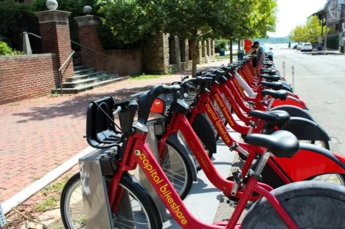 A row of red Capital Bikeshare bicycles are docked at a bike-sharing station on a sidewalk next to a street lined with trees and brick buildings, with a person in the background.