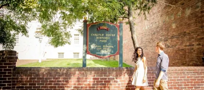 A smiling couple walks hand-in-hand past a sign for Carlyle House Historic Park, with brick walls, greenery, and sunlight in the background.