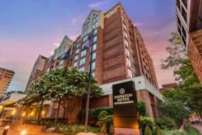 A modern, multi-story brick hotel building with a Sonesta ES Suites sign at the entrance, surrounded by trees, under a colorful evening sky.