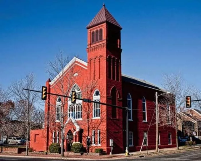 A historic red brick church with arched windows and a tall bell tower sits on a street corner under a clear blue sky, with bare trees and traffic lights in the foreground.