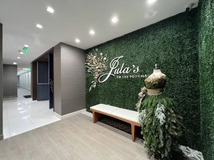A hallway with green artificial foliage walls featuring a sign that reads Julia’s on the Potomac, a white bench, and a mannequin dressed in a gown made of greenery and leaves.