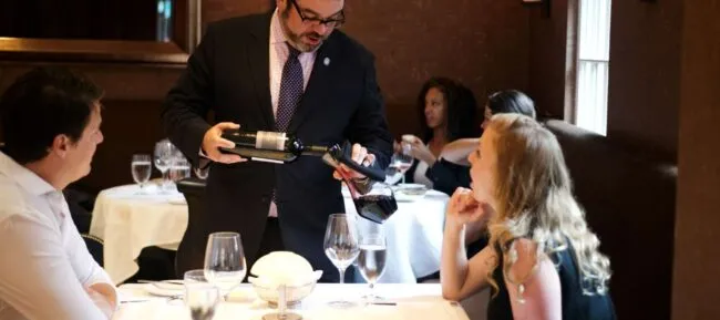 A sommelier in a suit pours red wine into a womans glass at an elegant restaurant table, while a man sits beside her. Other diners are visible in the softly lit background.
