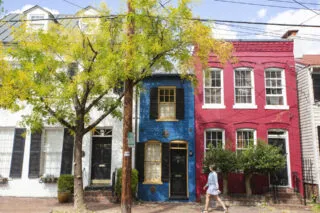 A person walks past three colorful row houses—white, blue, and red—on a tree-lined street with a sidewalk and utility pole in the foreground.