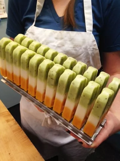 A person wearing a white apron and blue shirt holds a tray of neatly arranged tri-colored popsicles with green, white, and orange layers.