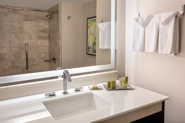A modern bathroom with a large mirror, white countertop with a sink, toiletries tray, and neatly folded towels hanging on the wall. A walk-in shower with a glass door and tiled walls is in the background.