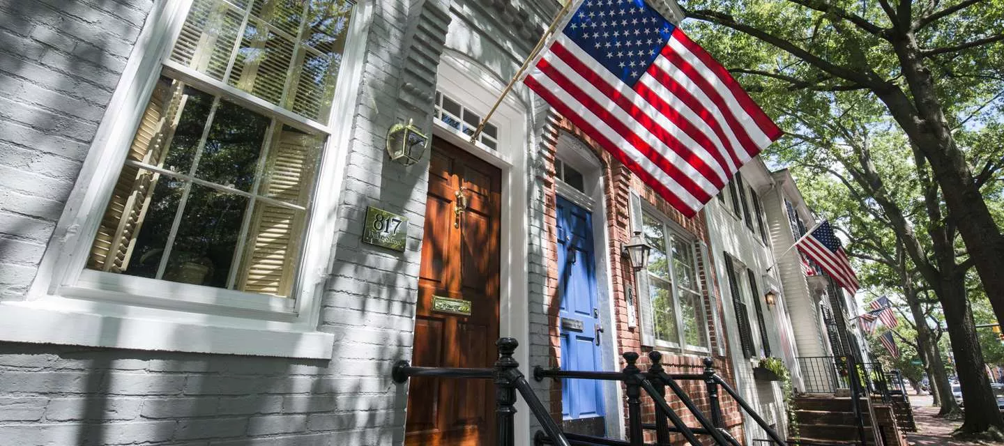A row of brick townhouses with American flags displayed, one with a brown door and another with a blue door, shaded by leafy trees on a sunny day.