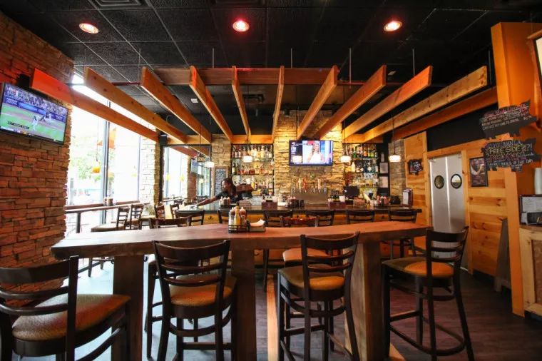 A cozy bar and restaurant with wooden tables and chairs, stone walls, a stocked bar, hanging beams on the ceiling, and several TVs showing sports above the bar and around the room.