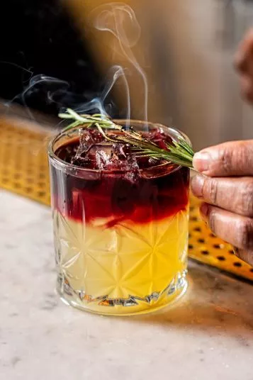 A hand garnishes a cocktail with a sprig of rosemary. The drink has a yellow base, a dark red layer on top, ice cubes, and is served in a patterned glass with wisps of smoke rising above it.
