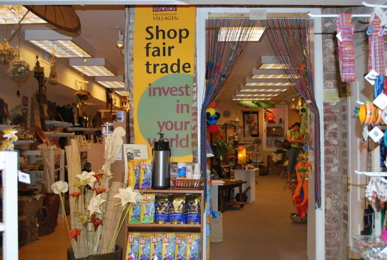 A store entrance displays a sign reading Shop fair trade, invest in your world. Inside, various colorful handmade goods, decorations, and crafts are visible, creating a vibrant and welcoming atmosphere.