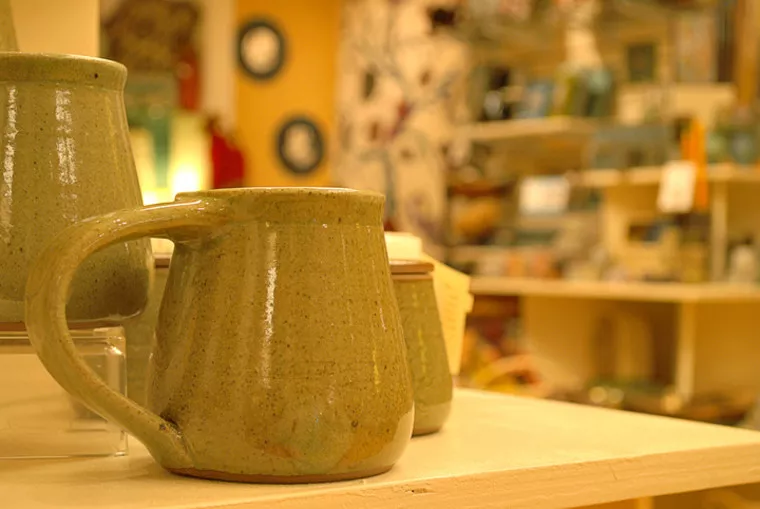 A close-up of a green ceramic mug with a handle on display in a pottery shop, surrounded by other pottery items on shelves in a warmly lit, blurred background.