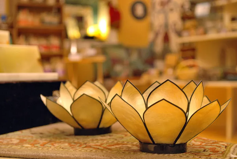 Two lotus-shaped candle holders with translucent, yellowish petals sit on a patterned surface, with a warmly lit, blurred background suggesting an indoor, cozy setting.