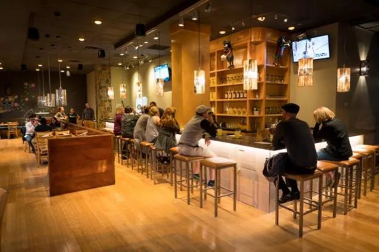 A modern bar with wooden floors and furniture, people sitting on stools at the counter, warmly lit pendant lights overhead, and several flat-screen TVs on the walls.