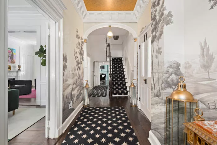A hallway with black star-patterned carpet, light-colored walls featuring landscape murals, arched doorway, brass lanterns on the right, and a view into a bright living room and a staircase at the end.