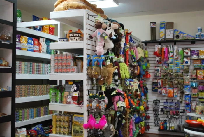Shelves in a pet store display plush animal toys, canned pet food, treats, and various pet accessories. The wall racks are filled with colorful toys and supplies for dogs and cats.