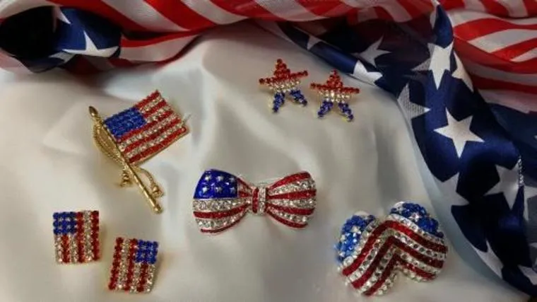 Patriotic jewelry with stars and stripes designs, including earrings, pins, and a bowtie, displayed on a white cloth next to an American flag with red, white, and blue colors.