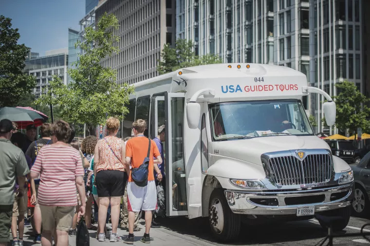 A group of people stand in line to board a white tour bus labeled USA Guided Tours on a sunny day in a city with modern glass buildings in the background.