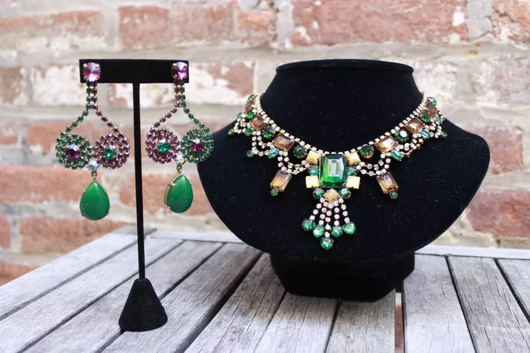 A sparkling jewelry set with green, gold, and purple gemstones includes a pair of dangling earrings and an ornate matching necklace, displayed on stands against a brick wall background.