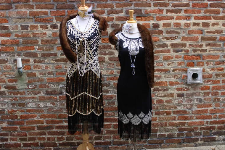 Two mannequins display vintage 1920s flapper dresses with beaded details, fringe, and fur stoles, set against a brick wall background.